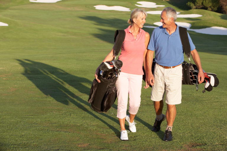 Senior Couple Walking Along Golf Course Carrying Bags