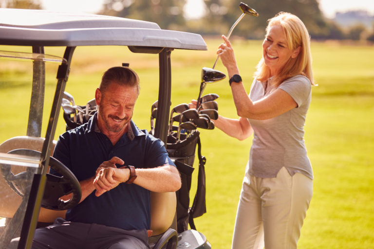 Man Checking Watch As Mature Couple Sit In Buggy And Play Round Of Golf Together